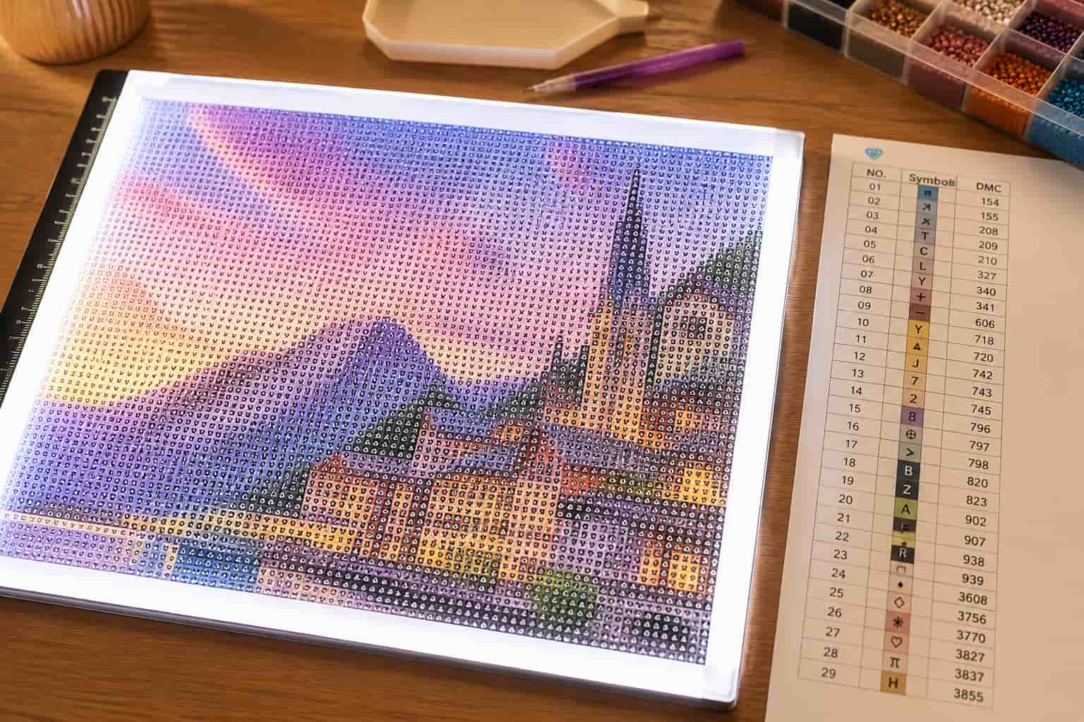 A completed diamond painting or cross-stitch style art piece depicting a scenic village with mountains and a church under a colorful sunset sky, displayed on a wooden table next to a color reference chart.