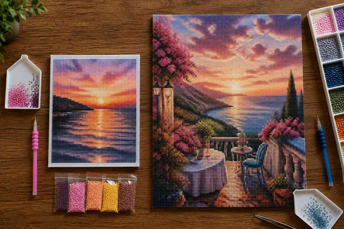 A diamond painting kit arranged on a wooden tabletop, featuring a completed scenic sunset artwork, a smaller preview of the same design, beads in pink, purple, orange, and yellow, a pink-handled needle tool, a blue-handled tool.