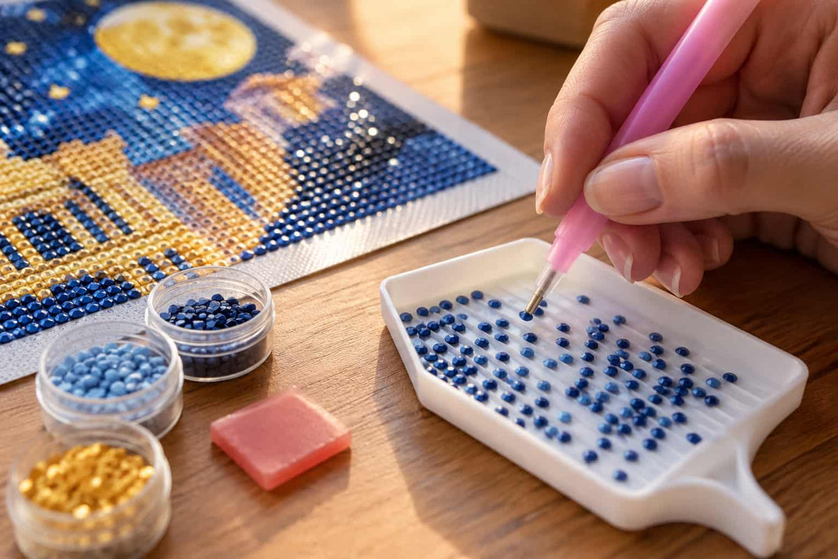 A close-up of a hand using a pink diamond painting pen to place blue beads into a white tray, with a partially completed night-sky canvas and small containers of blue and gold beads on a wooden table.