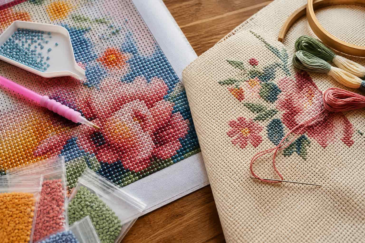 A close-up of a DIY diamond painting kit on a wooden table, showing a canvas with a floral design, beads, a pink-handled pick, and a finished embroidered fabric bag with matching thread and a wooden hoop.
