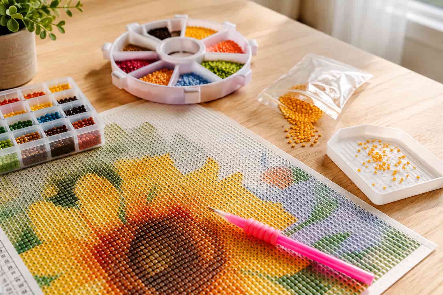 A diamond painting kit laid out on a wooden table, featuring a completed sunflower design, a pink stylus, a circular wheel of multicolor beads, a bead organizer tray, and small bags of loose beads.