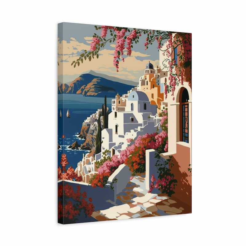 A rectangular canvas wall art print showing a Mediterranean coastal village scene with white buildings, pink flowering vines, blue sea, and mountains under a pale sky, photographed against a light gray background.