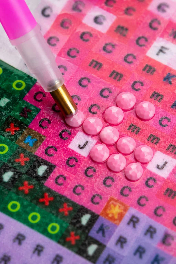 A close-up of a diamond painting or bead-on-board kit showing a pink pen-like tool applying faceted pink rhinestones to a printed pattern sheet with letters and colored symbols.