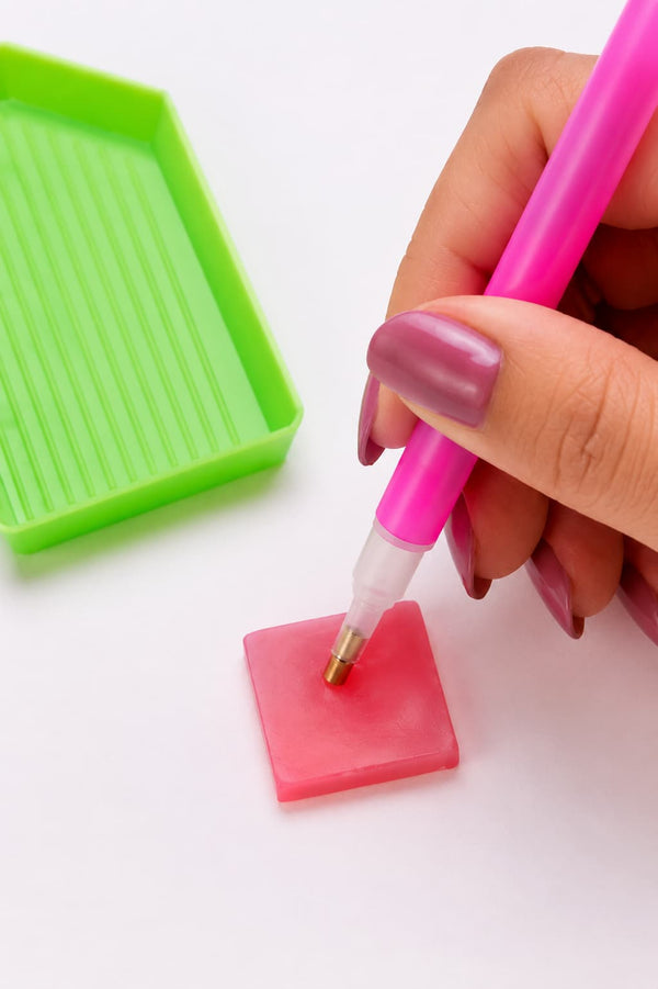 A close-up of a hand with purple manicured nails holding a pink pen-like tool over a small pink square, with a bright green rectangular tray in the background on a white surface.