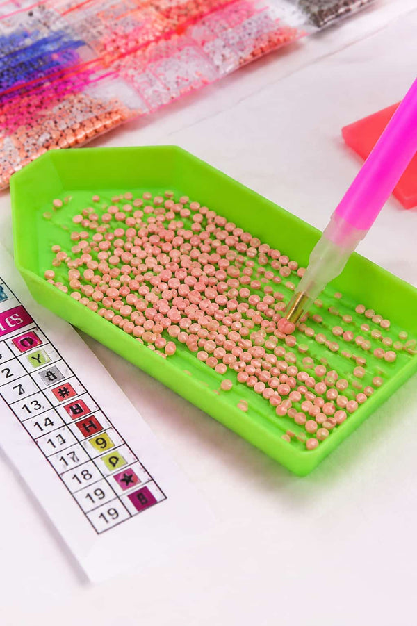 A close-up of a diamond painting or bead filling kit showing a green tray filled with small pink beads, a pink-handled applicator pen, and a color reference chart on a white surface.