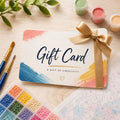 A gift card for creativity, styled on a light wood surface with paintbrushes, paint pots, and a coloring page. The card features a white background with colorful paint brushstrokes in blue, pink, and yellow, and is tied with a gold ribbon.