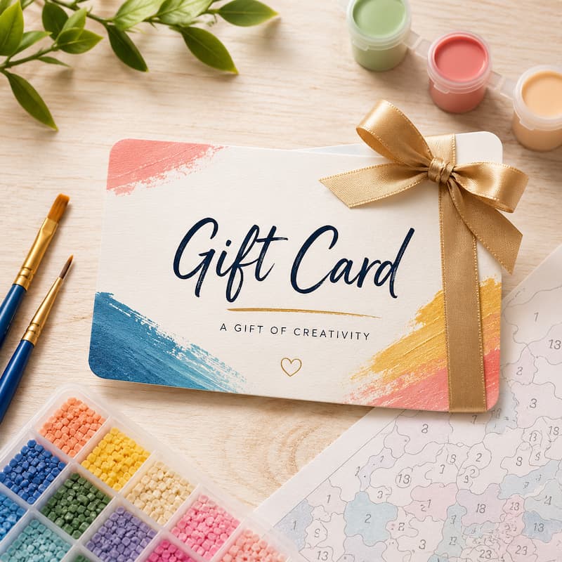 A gift card for creativity, styled on a light wood surface with paintbrushes, paint pots, and a coloring page. The card features a white background with colorful paint brushstrokes in blue, pink, and yellow, and is tied with a gold ribbon.