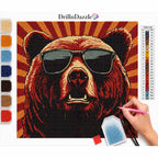 A diamond painting kit showing a completed bear portrait with sunglasses, a color list chart, a paint palette, and a hand holding a pink drill pen working on the project. The brand name 'DrillnDazzle' is visible at the top.