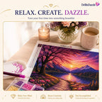 A DrillnDazzle diamond painting kit showing a completed landscape print of a sunset over a mountain lake, surrounded by craft supplies including a paint pen, color trays, and a mug on a wooden table.