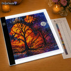 A diamond painting canvas showing an autumn forest scene with glowing orange trees and a purple night sky, displayed on a light box over a wooden table. A color list sheet is visible next to it, and candles and flowers decorate the background.
