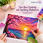 A person's hands are shown using a pink diamond painting pen to create a colorful cherry blossom sunset scene on a canvas. The scene is styled as a flat-lay with a marble background, a lit candle, a potted plant, and a box of colorful diamond painting beads.