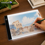 An illuminated drawing tablet on a wooden desk, showing a painted scene of buildings and sky. A hand holds a green pencil over the screen, with a plant and notebook in the background.