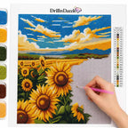 A person's hand holding a pink paint pen is painting on a canvas next to a completed diamond painting of a sunflower field under a blue sky, with color swatches and a color list visible on the left and right.